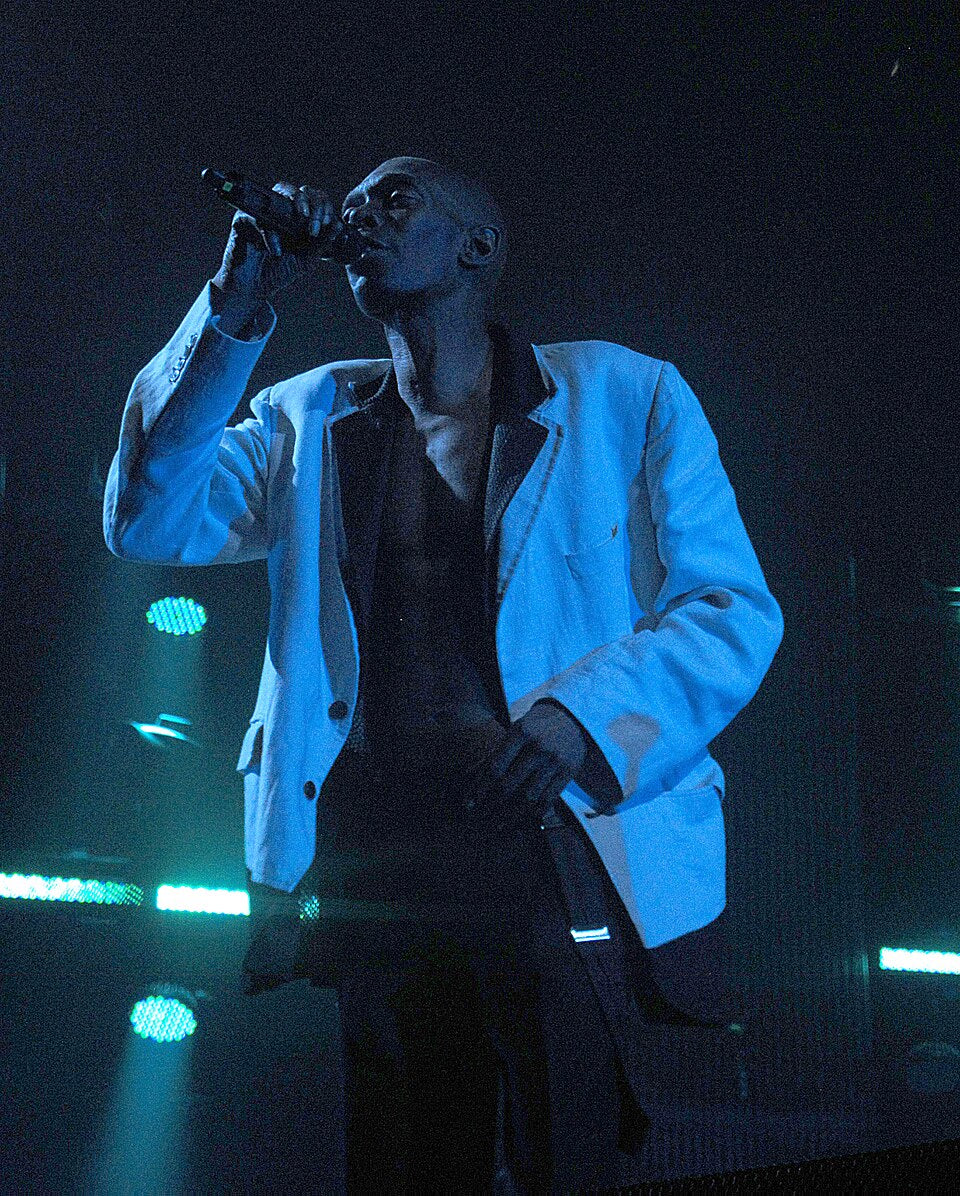 Maxi Jazz zingt met een microfoon in zijn hand, in een witte jas onder blauw podiumlicht.