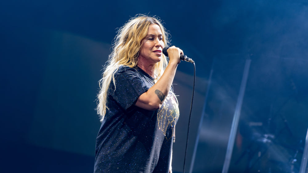 Alanis Morissette zingt met een handmicrofoon op een blauw verlicht podium.