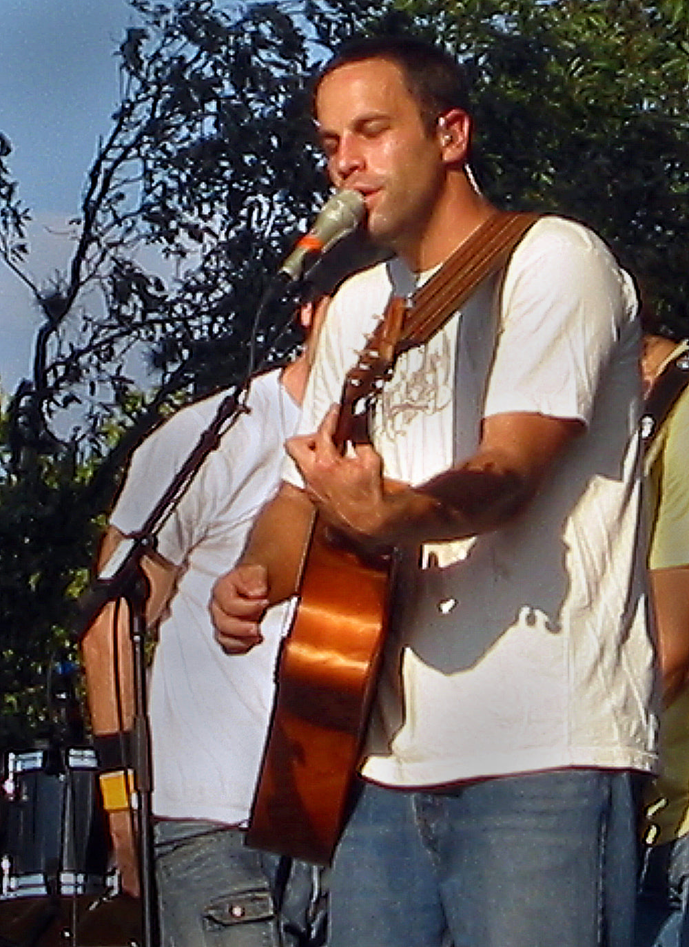 Jack Johnson zingt en speelt akoestische gitaar op een buitenpodium, met bomen op de achtergrond.