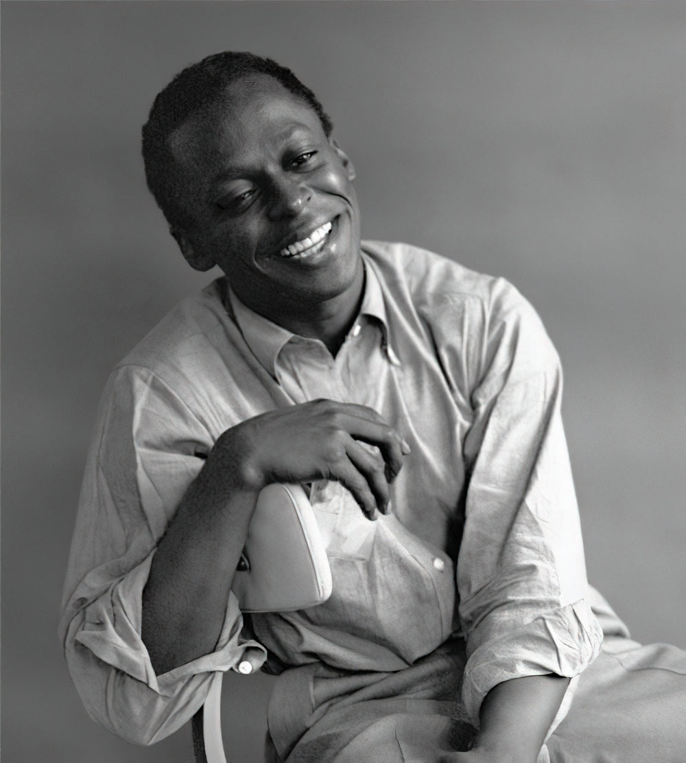Miles Davis glimlacht en leunt met zijn arm op de rug van een stoel.