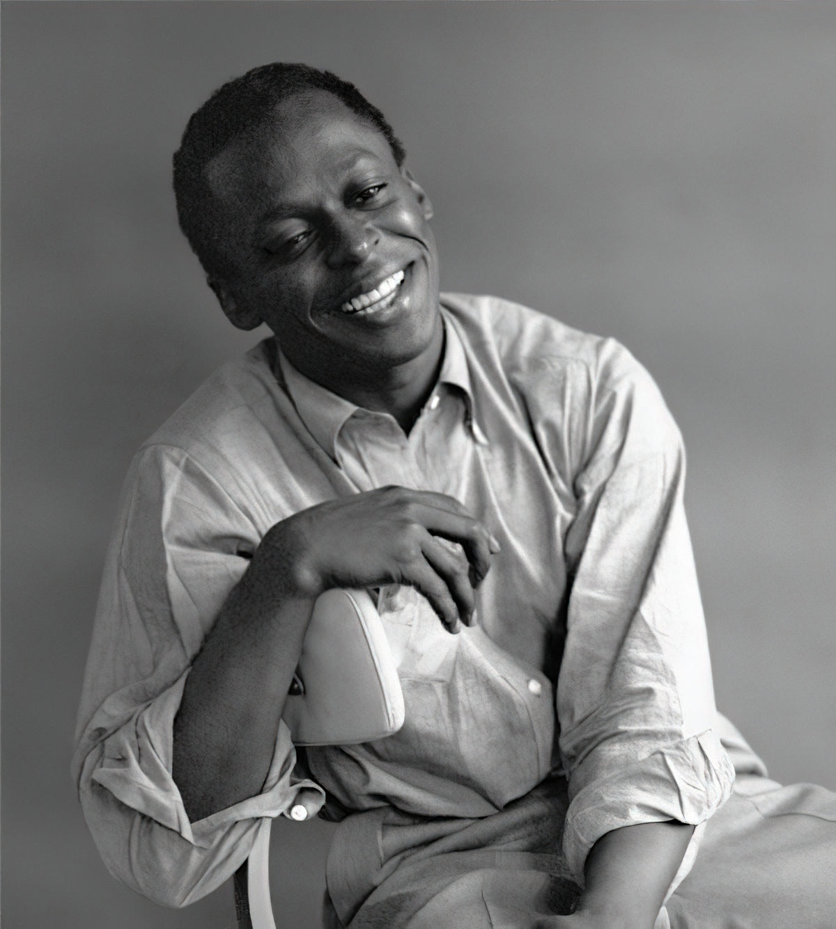 Miles Davis glimlacht en leunt met zijn arm op de rug van een stoel.
