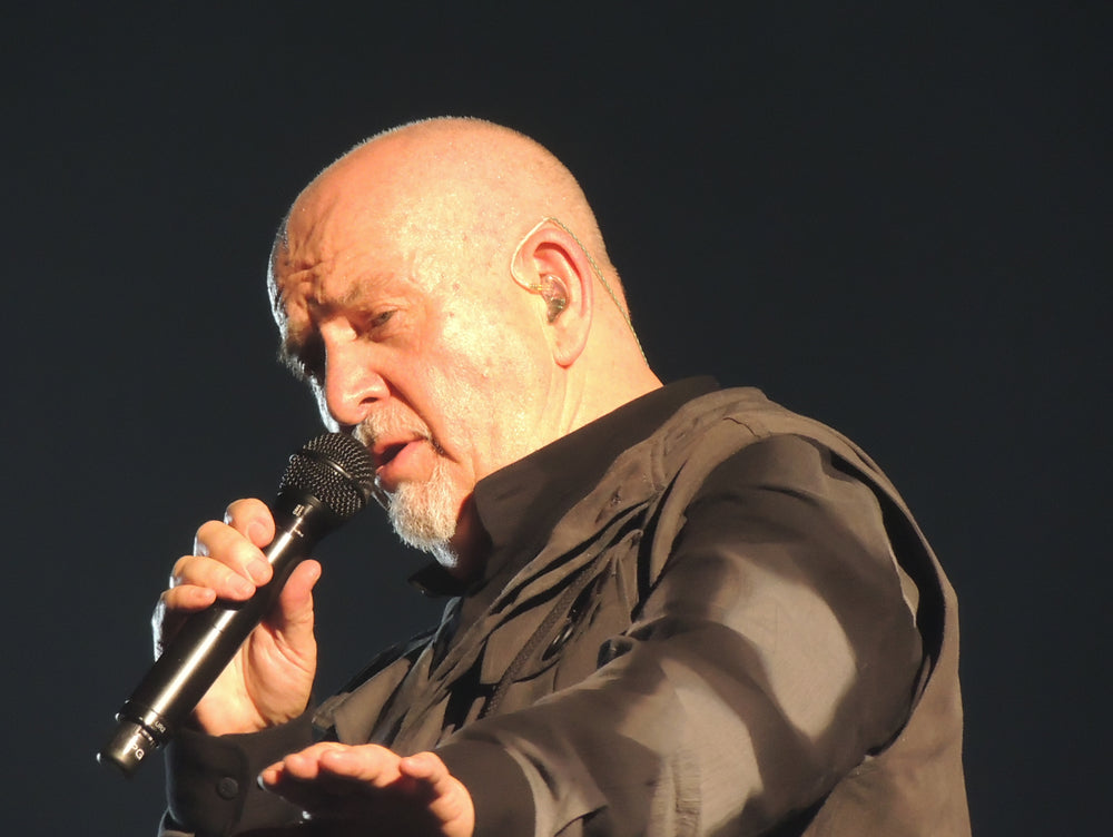 Peter Gabriel zingt in een handmicrofoon en strekt zijn vrije hand uit naar het publiek.