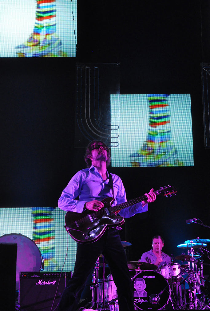 Jarvis Cocker van Pulp speelt gitaar tijdens Coachella op 20 april 2012, verlicht door paars podiumlicht.