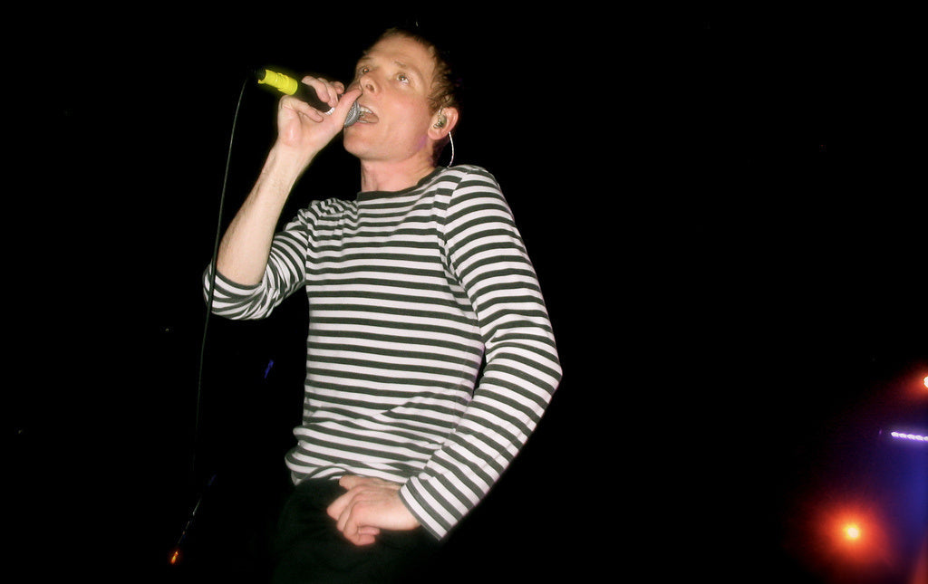 Stuart Murdoch van Belle & Sebastian zingt met een handmicrofoon, in een gestreept shirt op een donker podium.