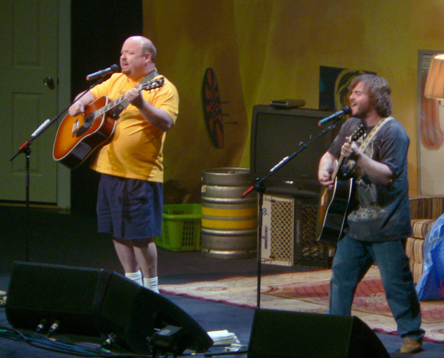 Jack Black en Kyle Gass van Tenacious D met akoestische gitaren op een podium ingericht als woonkamer