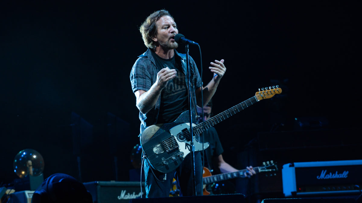 Eddie Vedder zingt met een gitaar om zijn schouder op een blauw verlicht podium.
