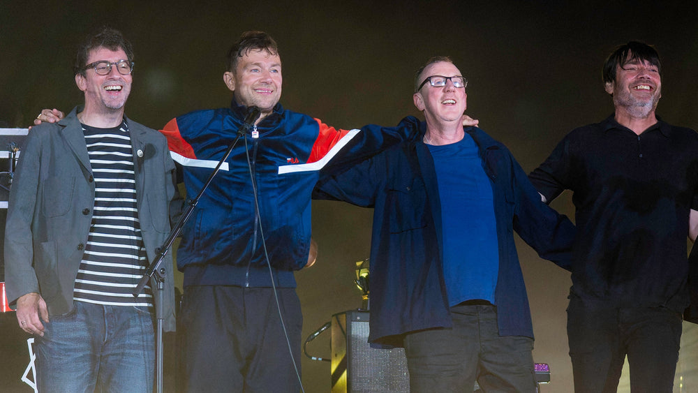 Graham Coxon, Damon Albarn, Dave Rowntree en Alex James staan lachend naast elkaar op het podium, met hun armen om elkaar.