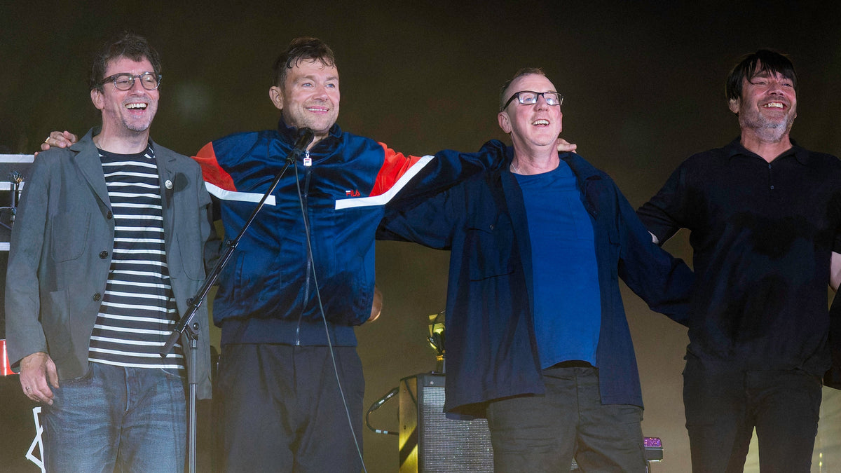Graham Coxon, Damon Albarn, Dave Rowntree en Alex James staan lachend naast elkaar op het podium, met hun armen om elkaar.