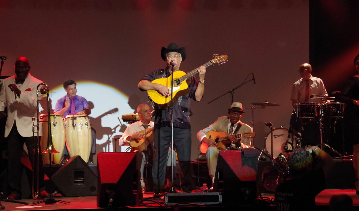 Eliades Ochoa zingt en speelt gitaar met de Orquesta Buena Vista Social Club op een podium.