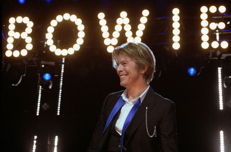 David Bowie glimlacht op het podium, met zijn naam in lichtletters achter zich.
