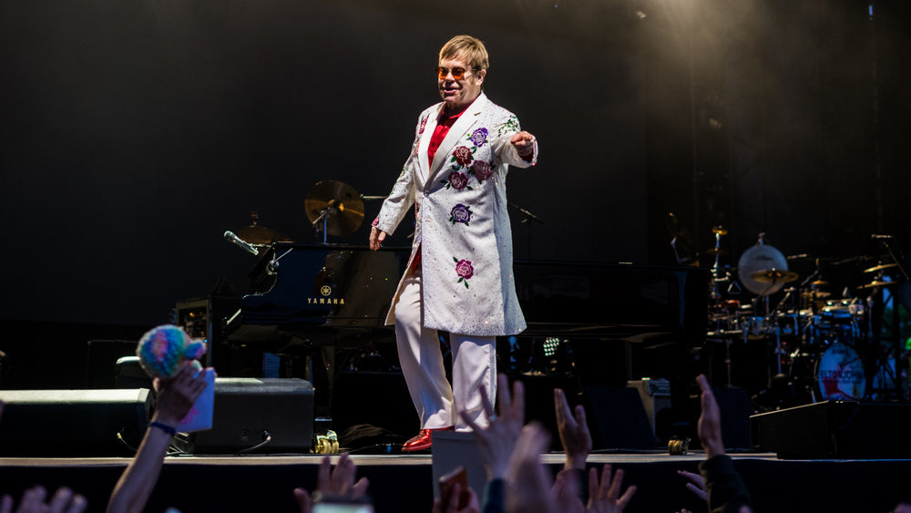 Elton John in een licht kostuum met geborduurde bloemen, staand voor zijn piano op het podium.