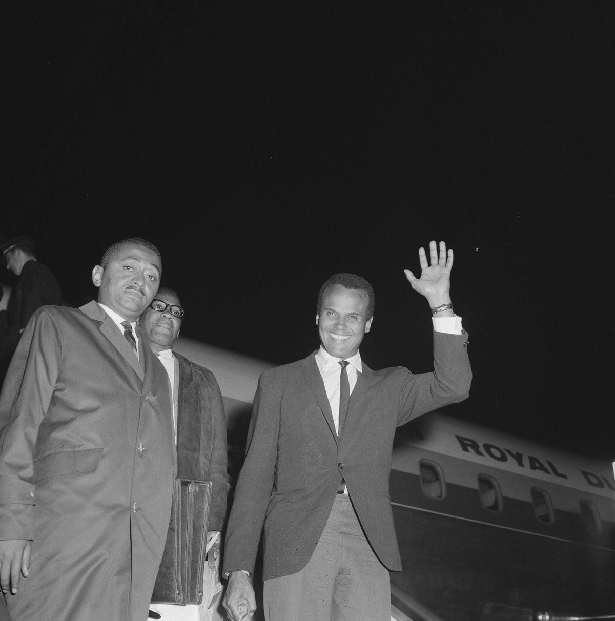 Harry Belafonte zwaait naast twee mannen op Schiphol, met een vliegtuig op de achtergrond.