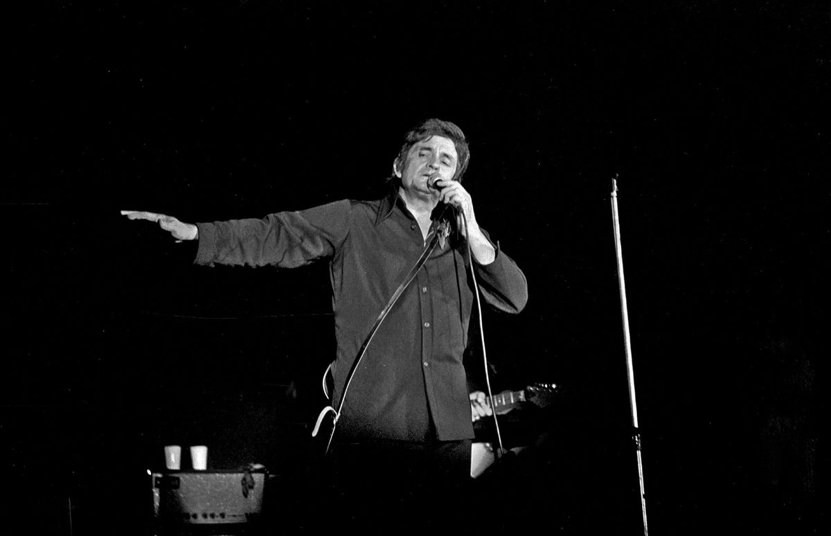 Johnny Cash zingt in een microfoon met een arm uitgestrekt op een donker podium.