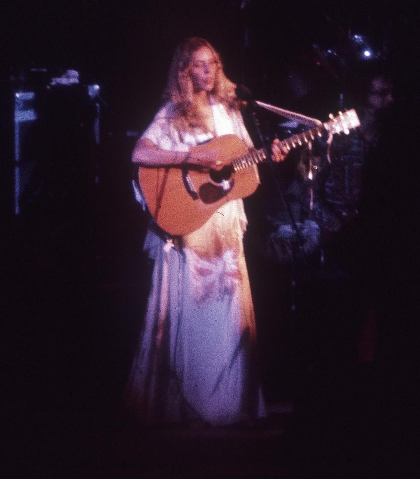 Joni Mitchell zingt en speelt akoestische gitaar op een podium in 1974.