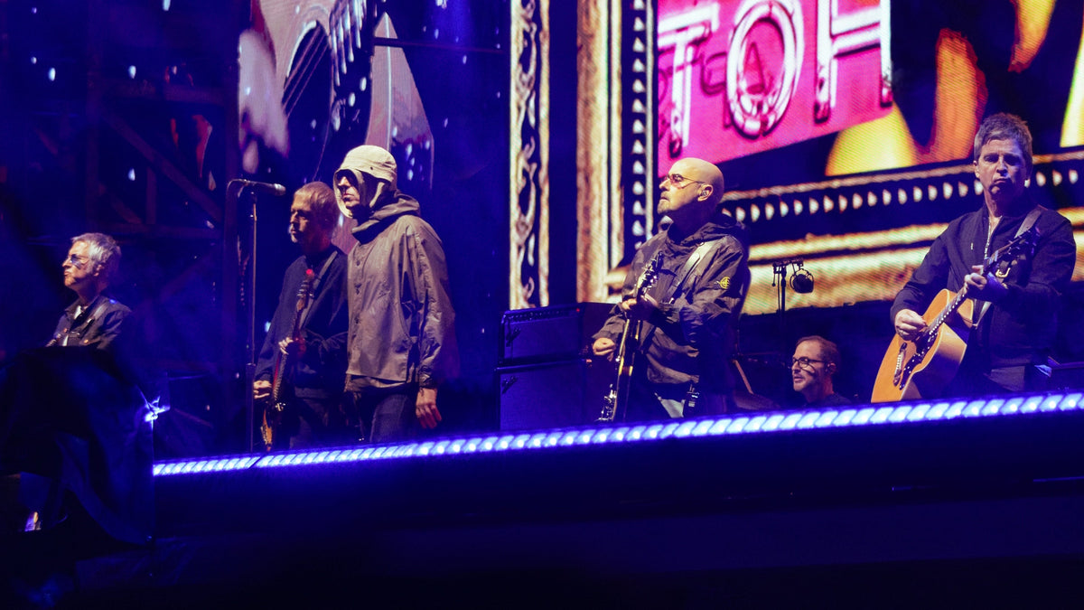 Oasis op het podium van Wembley, met Liam Gallagher bij de microfoon en Noel Gallagher rechts met gitaar.