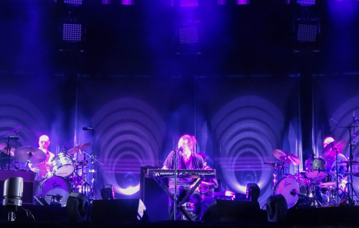 Thom Yorke achter een keyboard, tussen twee drumstellen en paars podiumlicht tijdens Radiohead in 2016