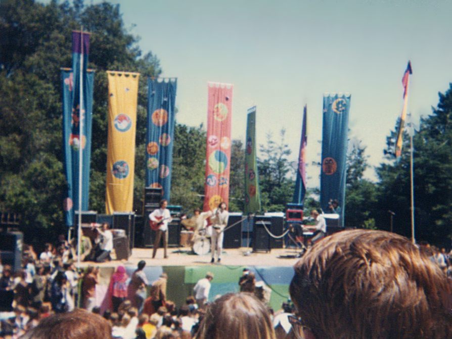 The Doors spelen op een buitenpodium tussen hoge gekleurde vlaggen, gezien vanuit het festivalpubliek.