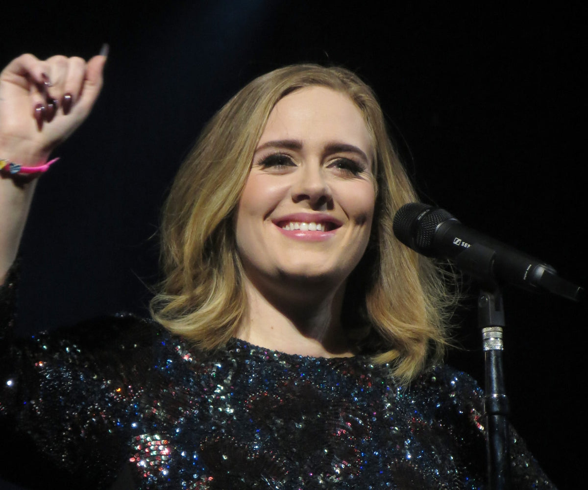 Adele glimlacht naast een microfoon en steekt haar rechterhand op tijdens een optreden in Glasgow in 2016, in een zwarte jurk met glinsterende pailletten.