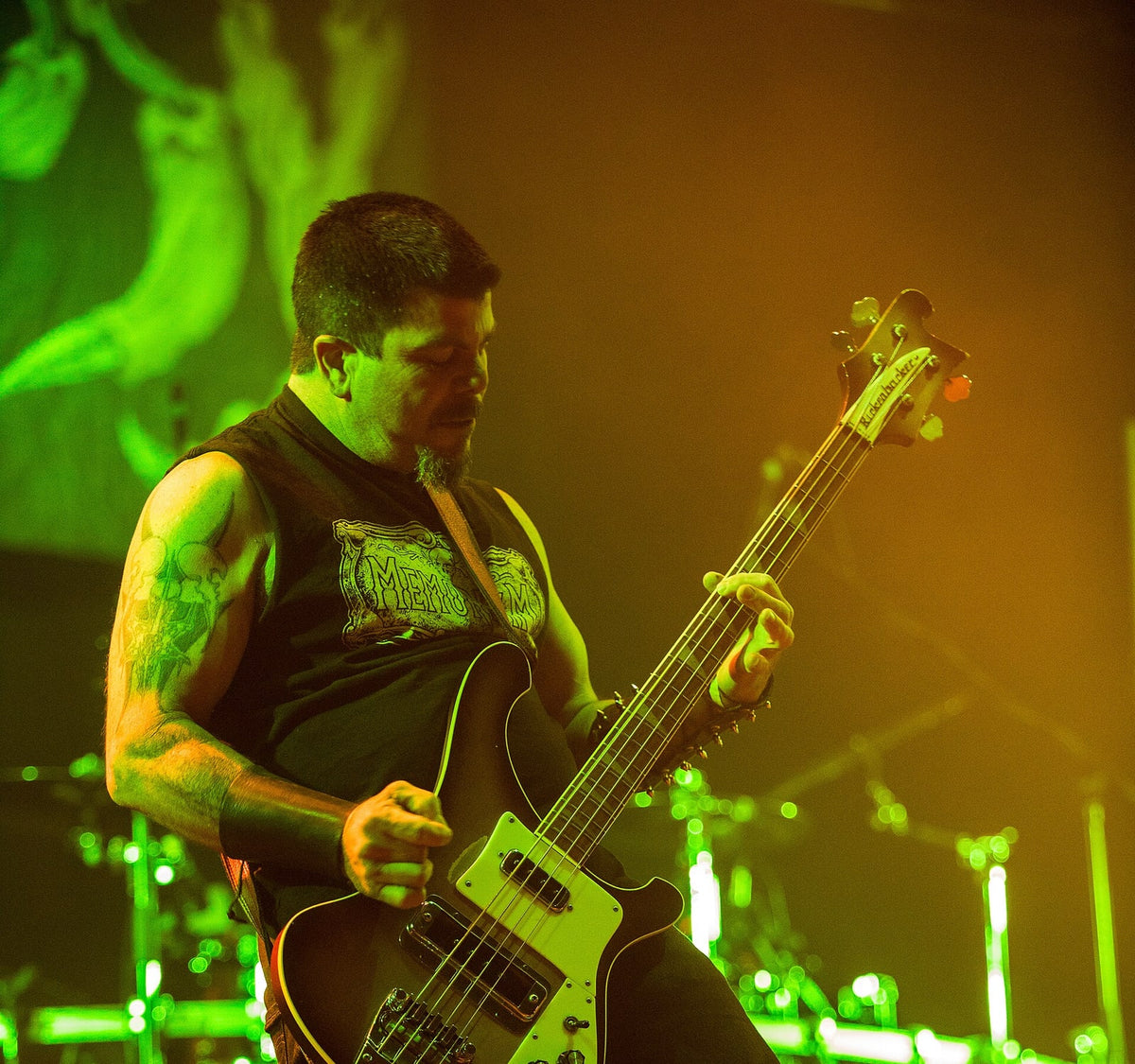 De bassist van Autopsy tijdens Party.San Metal Open Air 2017, met kort donker haar, een sik en getatoeëerde armen, spelend op een basgitaar in groen-geel podiumlicht.