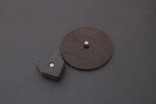 Zwart vierkant blokje met een zilveren pin en een bruin rond stuk schuurpapier met een pin.
