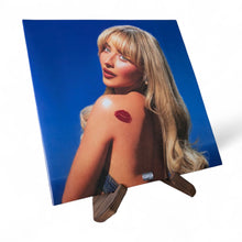 Albumcover met blondharige vrouw met rode lippenafdruk op haar schouder, tegen een blauwe achtergrond.